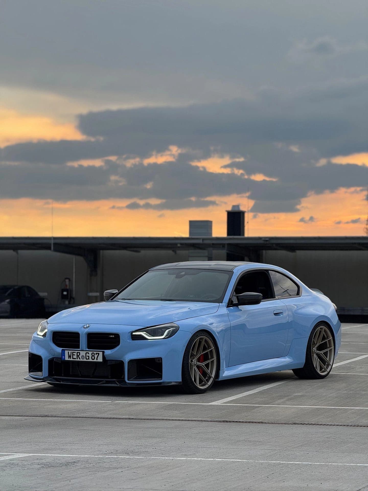 Hellblauer BMW M2 mit bronzefarbenen Felgen auf dem Parkdeck im Abendlicht